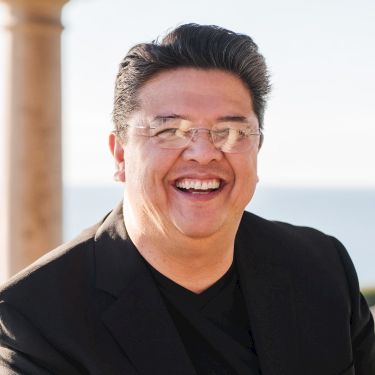 A cheerful man in a black blazer and glasses smiles warmly outdoors by the water, with columns in the background.