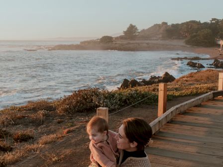 A person sits on a wooden boardwalk by the coast, holding a baby, with the ocean and rocky shore in the background at sunset.