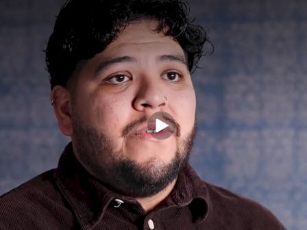 Close-up portrait of a bearded man with curly hair against a bluish background, with a video play icon across his mouth.