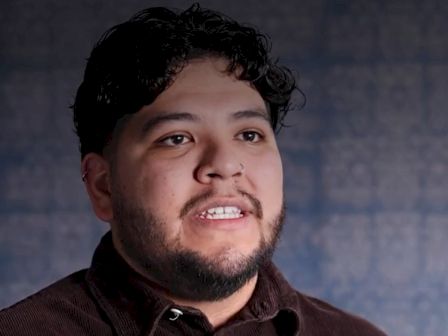 A close-up portrait of a young man with dark hair and beard speaking against a blurred blue background.