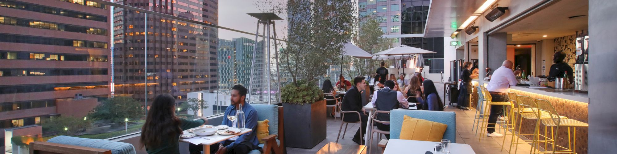 Rooftop patio dining with people, a warm fire pit, and a skyline of skyscrapers labeled First Republic and CTBC Bank at dusk.